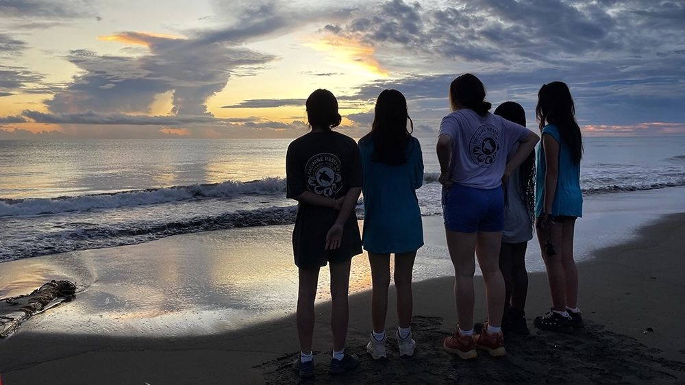 Five people stand on a beach at sunset, facing the ocean. The sky is partly cloudy with orange and blue hues reflected on the wet sand. The group appears to be watching the horizon peacefully.