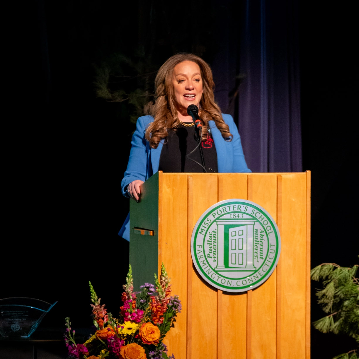 A woman in a blue blazer speaks at a wooden podium with the Miss Porters School seal. Colorful flowers are arranged in front of the podium on a stage with dark curtains in the background.