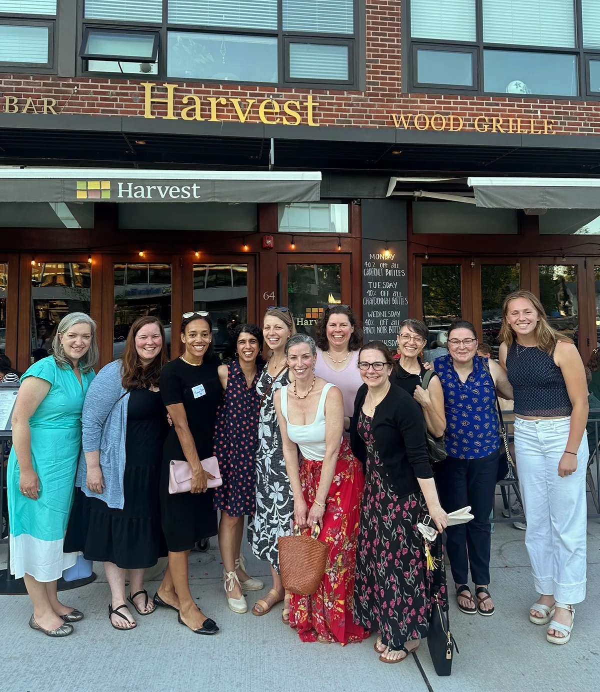 A group of twelve women smile and pose together outside Harvest restaurant, dressed in summer outfits, standing on a sidewalk in front of the brick building.
