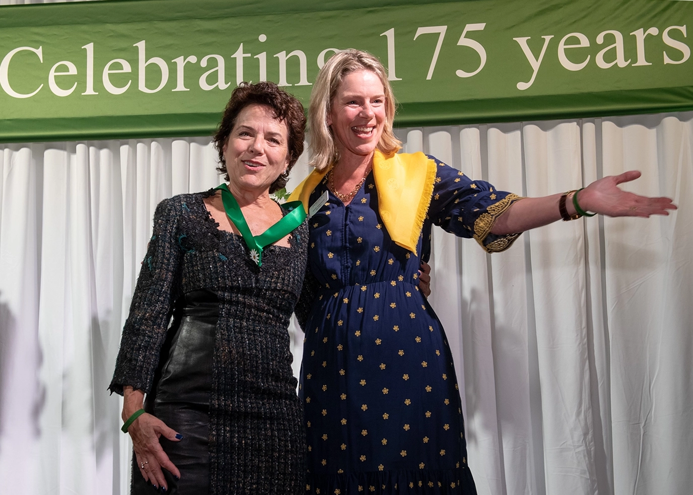 Two women stand smiling together in front of a green banner that reads Celebrating 175 years. One woman gestures with her arm, and both wear dresses and green sashes or accessories. White curtains hang behind them.