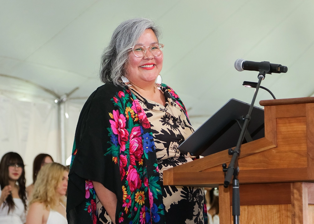 A smiling woman with gray hair, glasses, and a colorful floral shawl stands at a podium with a microphone, holding a folder. People are visible in the background under a white tent.