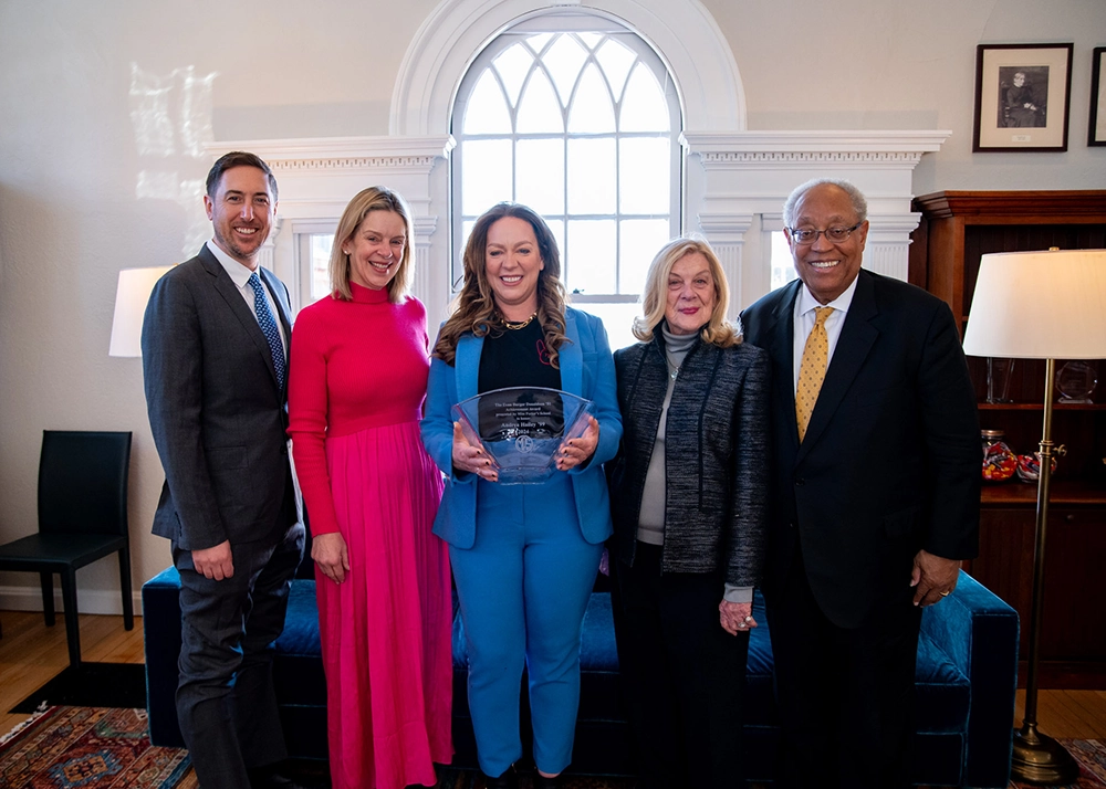 Five people stand indoors, the person in the center holds a clear glass award. All are smiling, dressed in business or formal attire, with a large arched window and framed photos in the background.