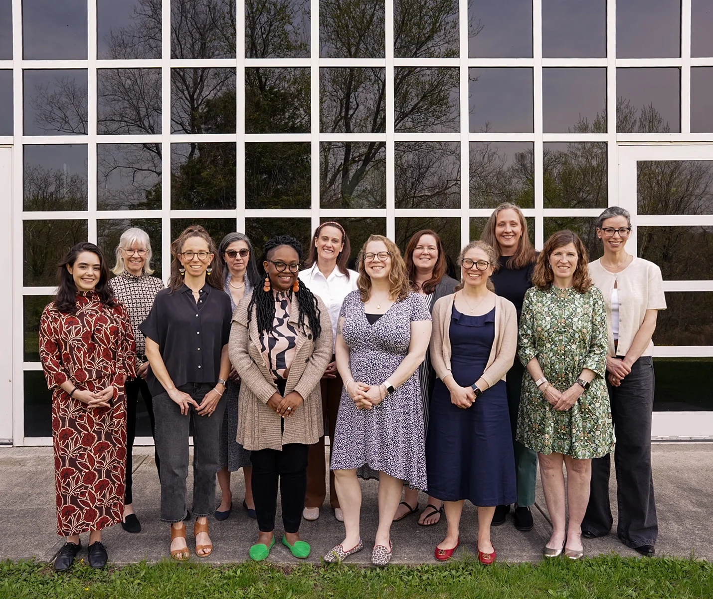 Thirteen women stand in two rows, smiling, in front of a large grid of windows with green trees reflected behind them. They are dressed in a variety of casual and professional outfits.