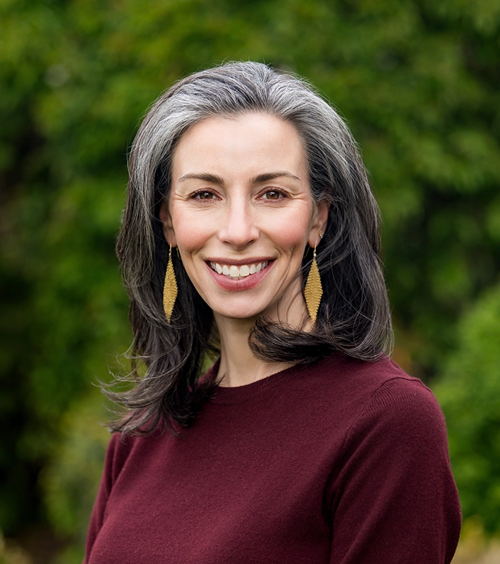A woman with long, wavy gray and brown hair, wearing a burgundy top and gold leaf-shaped earrings, smiles outdoors with green foliage in the background.