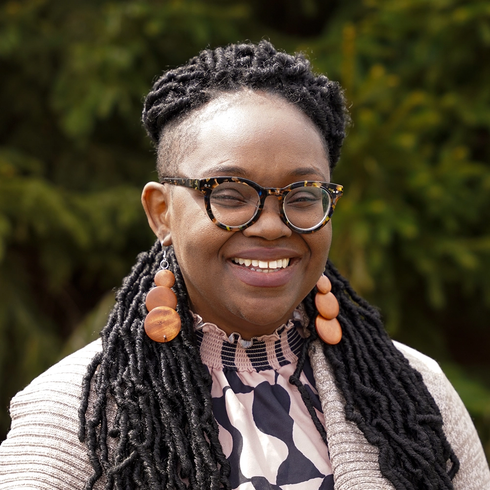 A woman with glasses and long braids smiles outdoors, wearing large round earrings and a patterned blouse with a light cardigan. Green foliage is blurred in the background.