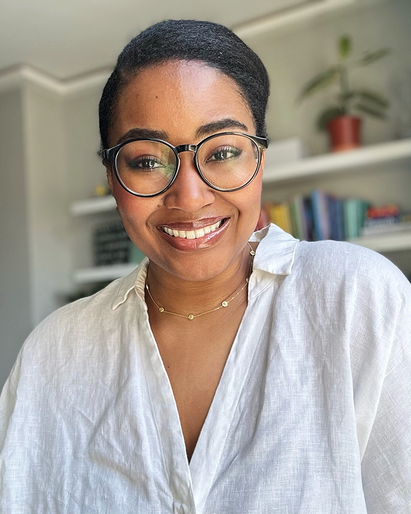 A woman with glasses and a white shirt smiles at the camera. She has a short hairstyle and is wearing a gold necklace. Books and a potted plant are visible on shelves in the background.