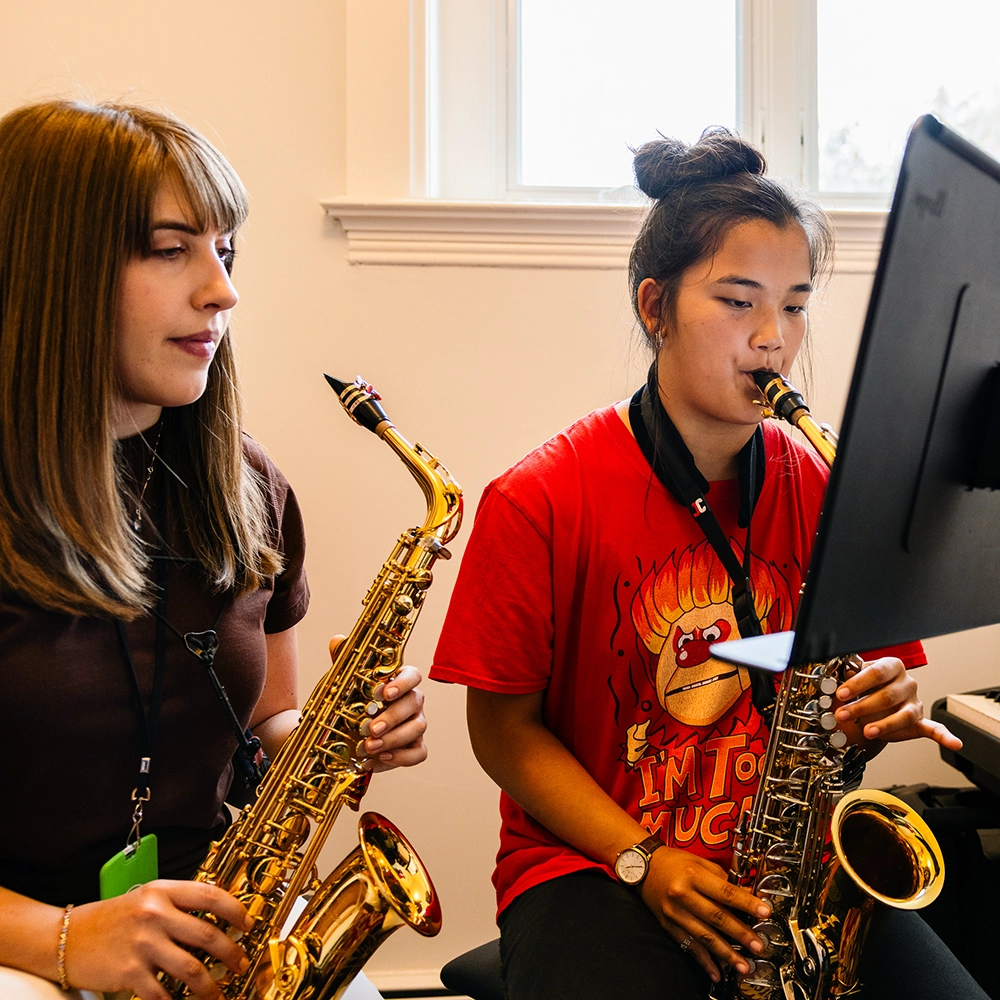 Two young women play saxophones while looking at a monitor. One wears a red shirt with a cartoon graphic, the other a brown shirt. They are seated in a bright room with a window in the background.
