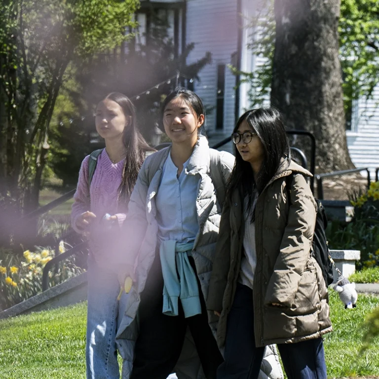 Three young women, wearing jackets and carrying backpacks, walk together outside on a sunny day, with green grass, trees, and flowers visible in the background.
