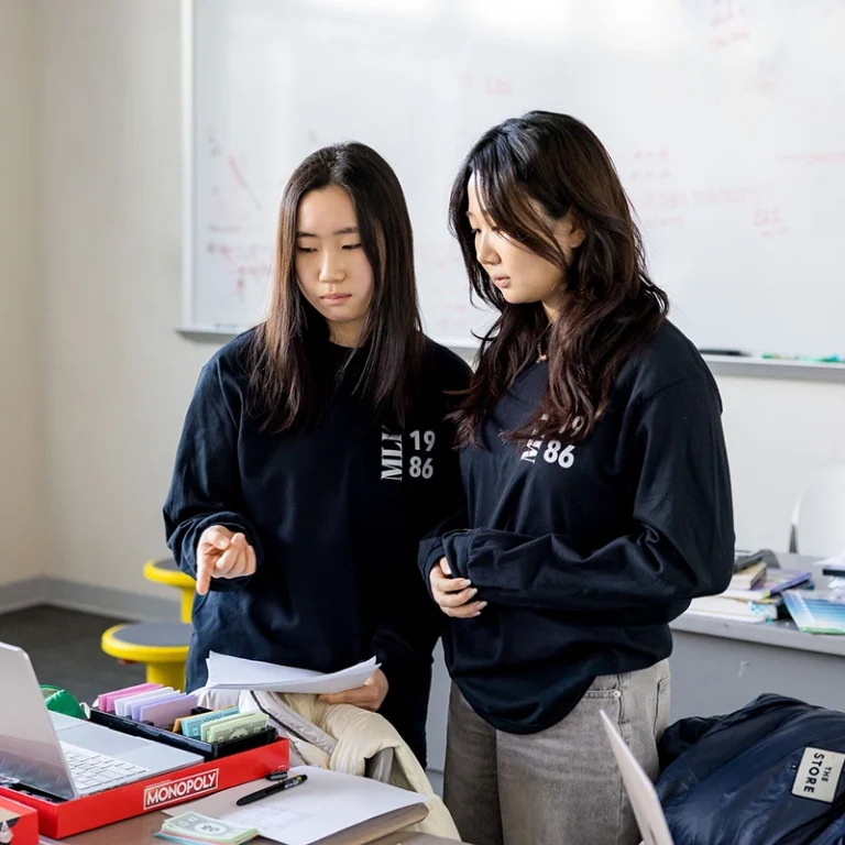 Two women in black shirts stand by a desk with Monopoly money, papers, and a laptop. They appear focused, possibly discussing work or studying, with a whiteboard covered in notes behind them.