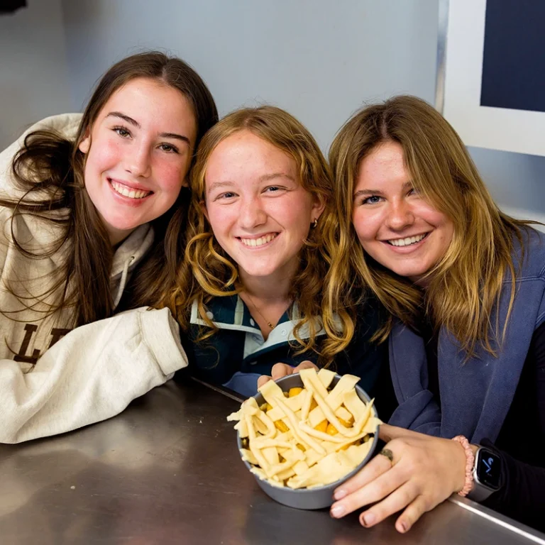 Three smiling young women sit at a table with a bowl of uncooked fries in front of them. They lean in close to each other, posing happily for the photo.
