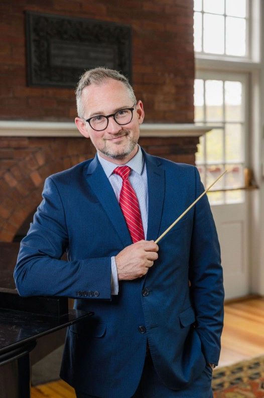 MPS Music Director Patrick Reardon stands indoors, holding a conductors baton and smiling, with a brick fireplace and large windows in the background.