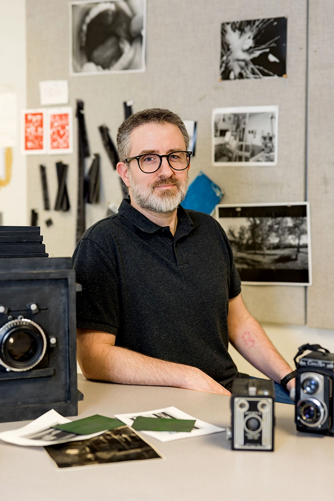 Photography teacher Josh Bezdek in the film photography studio with vintage film cameras on the table and negatives and photo prints on the wall