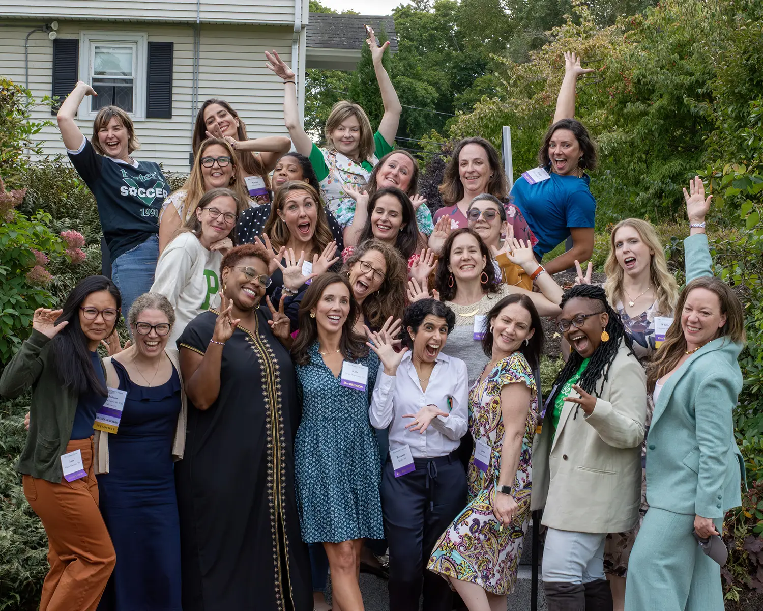 A group of women pose outdoors, smiling and making playful hand gestures. Most are wearing conference badges. Trees and a house are visible in the background.