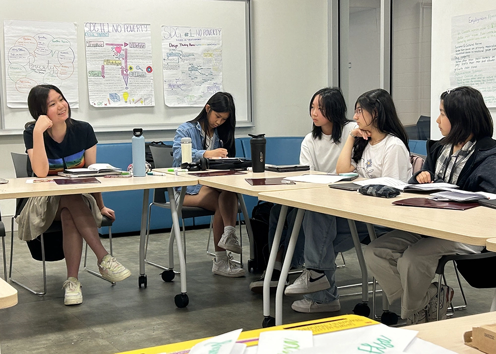Five students sit around tables in a classroom, engaged in discussion. Colorful posters with handwritten notes are displayed on the wall behind them.