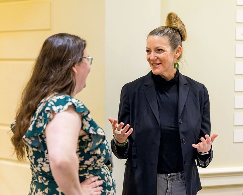 Two women stand indoors having a friendly conversation. One woman wears a floral dress and glasses, while the other wears a black blazer and a topknot hairstyle. Both appear engaged and expressive.