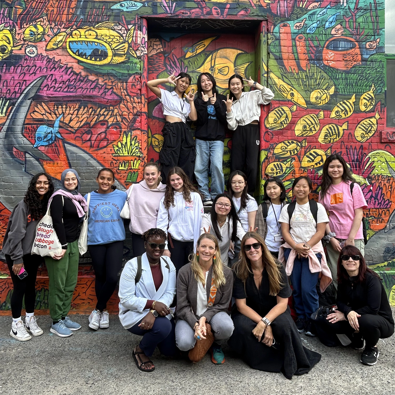 A group of people pose and smile in front of a colorful wall mural featuring animals, plants, and vibrant designs. The group is diverse in age and style, with some sitting and others standing or kneeling.