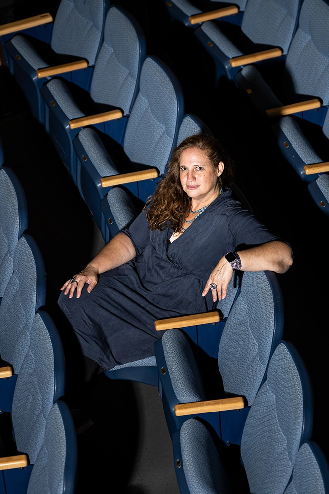 MPS theater director Alexandra London-Thompson sits alone in a row of blue theater seats, wearing a dark dress and looking at the camera with a slight smile. The lighting highlights her in the mostly empty auditorium.