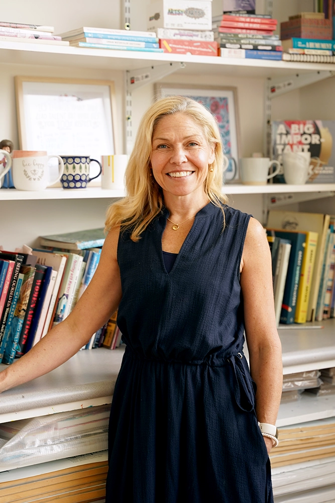 A woman with blonde hair in a navy dress stands smiling in front of bookshelves filled with books, mugs, and framed art in a bright room.