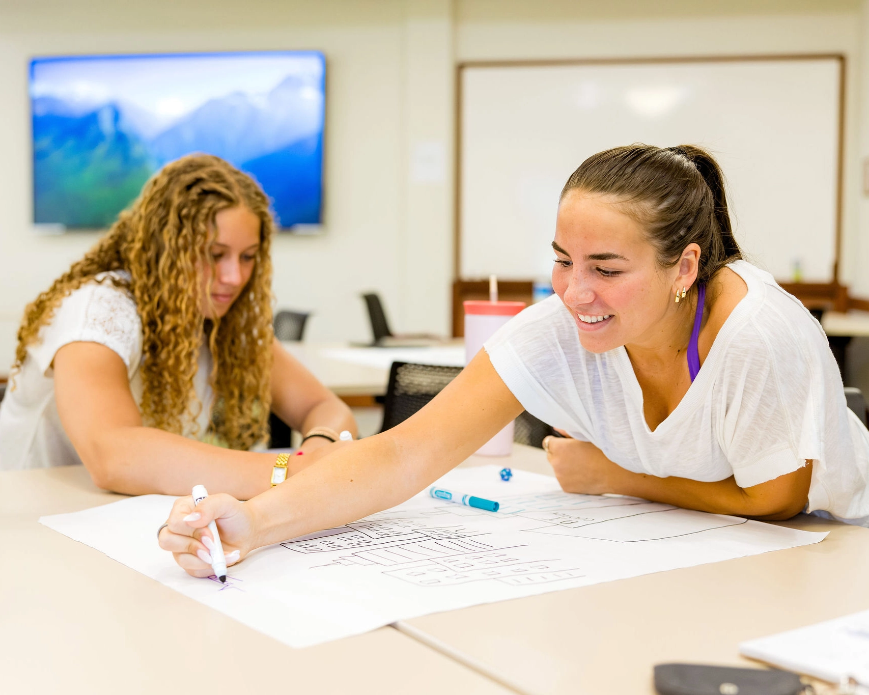Two students sit at a table in a classroom, working together on a large sheet of paper. One student smiles while writing with a marker, and the other focuses on the paper. A large monitor and whiteboard are in the background.