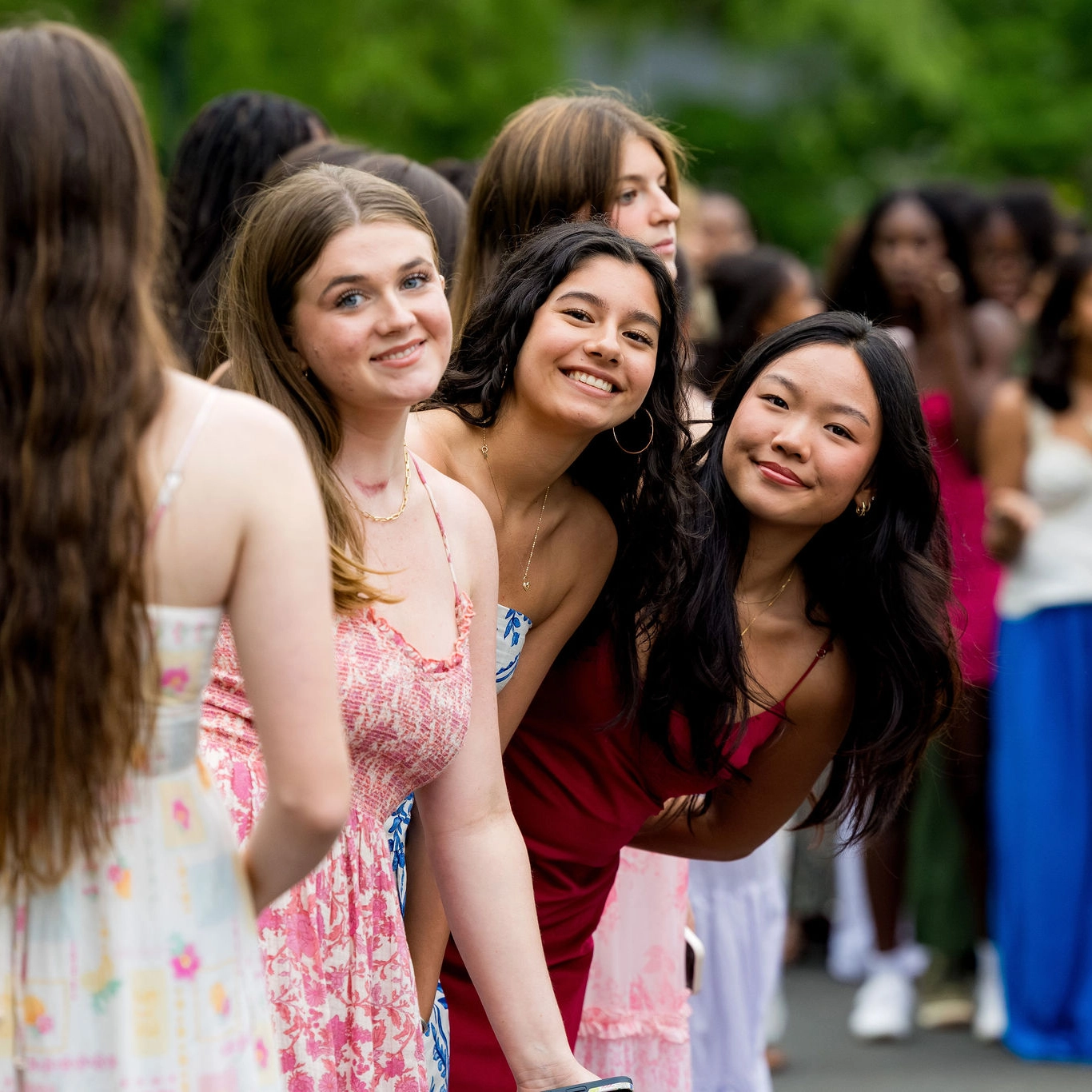 A group of young women in colorful dresses stand outside, smiling and posing together. The focus is on three who lean forward, looking at the camera, with others blurred in the background.