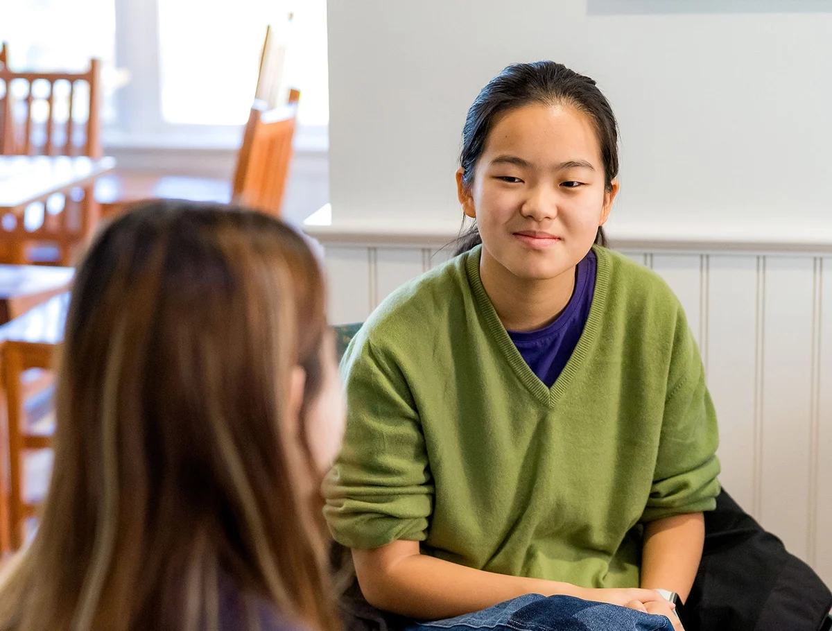 Two people sit at a table. One faces the camera, wearing a green sweater, and the other has their back turned, holding a partially eaten donut. There are pastries and drinks on the table.
