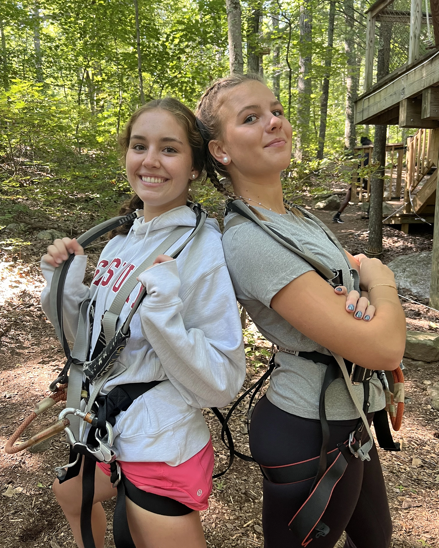 Two young women stand back-to-back in a forest, smiling and wearing harnesses, likely for a ropes course or zip-lining. They appear confident and ready for adventure, with trees and a wooden structure in the background.