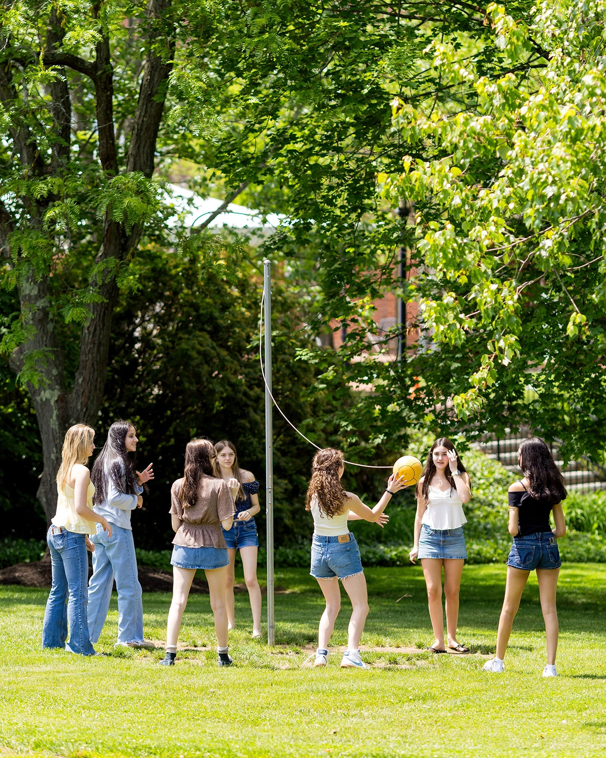 Seven teenage girls are playing tetherball outside on a sunny day. They are standing on grass under leafy trees, with one girl hitting the yellow ball attached to a pole.