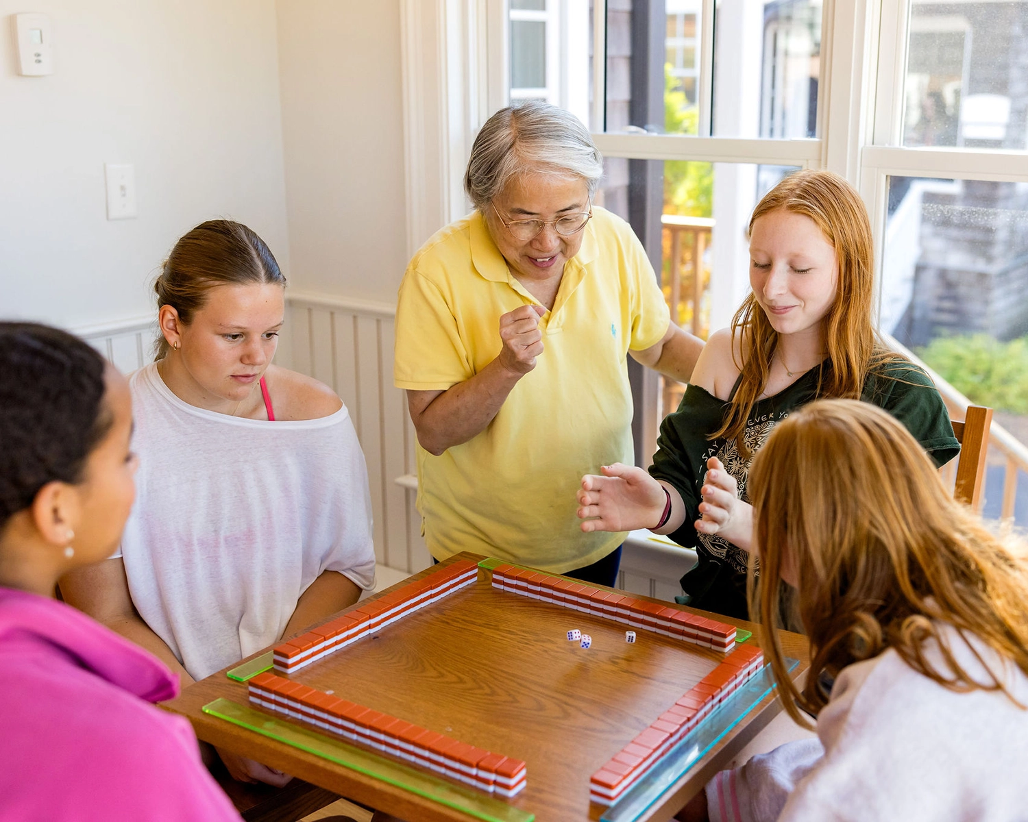 Four people are sitting around a table playing a dice game, while an older person in a yellow shirt stands and watches, smiling. Bright daylight streams through large windows behind them.