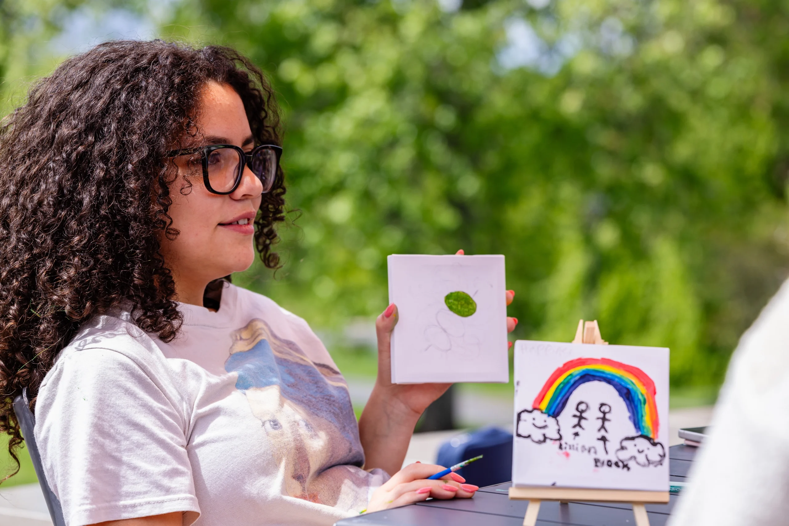 A person with curly hair and glasses holds up a drawing. Another colorful painting with a rainbow, clouds, and stick figures is displayed on a small easel. Green trees blur in the background.