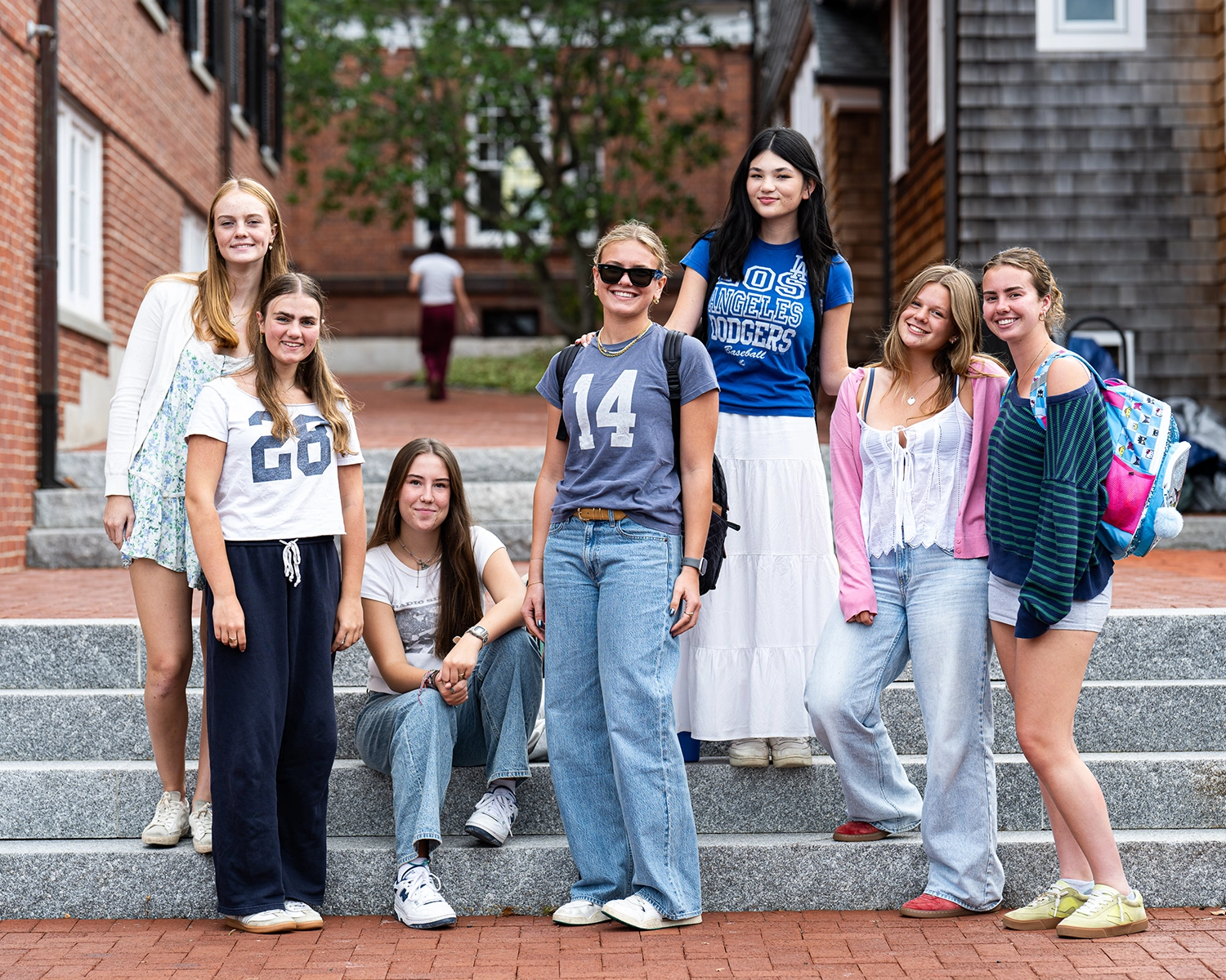 Seven young women pose and smile on outdoor steps in front of brick buildings. Most wear casual clothes such as jeans and T-shirts. The group appears relaxed and happy, enjoying a bright day.