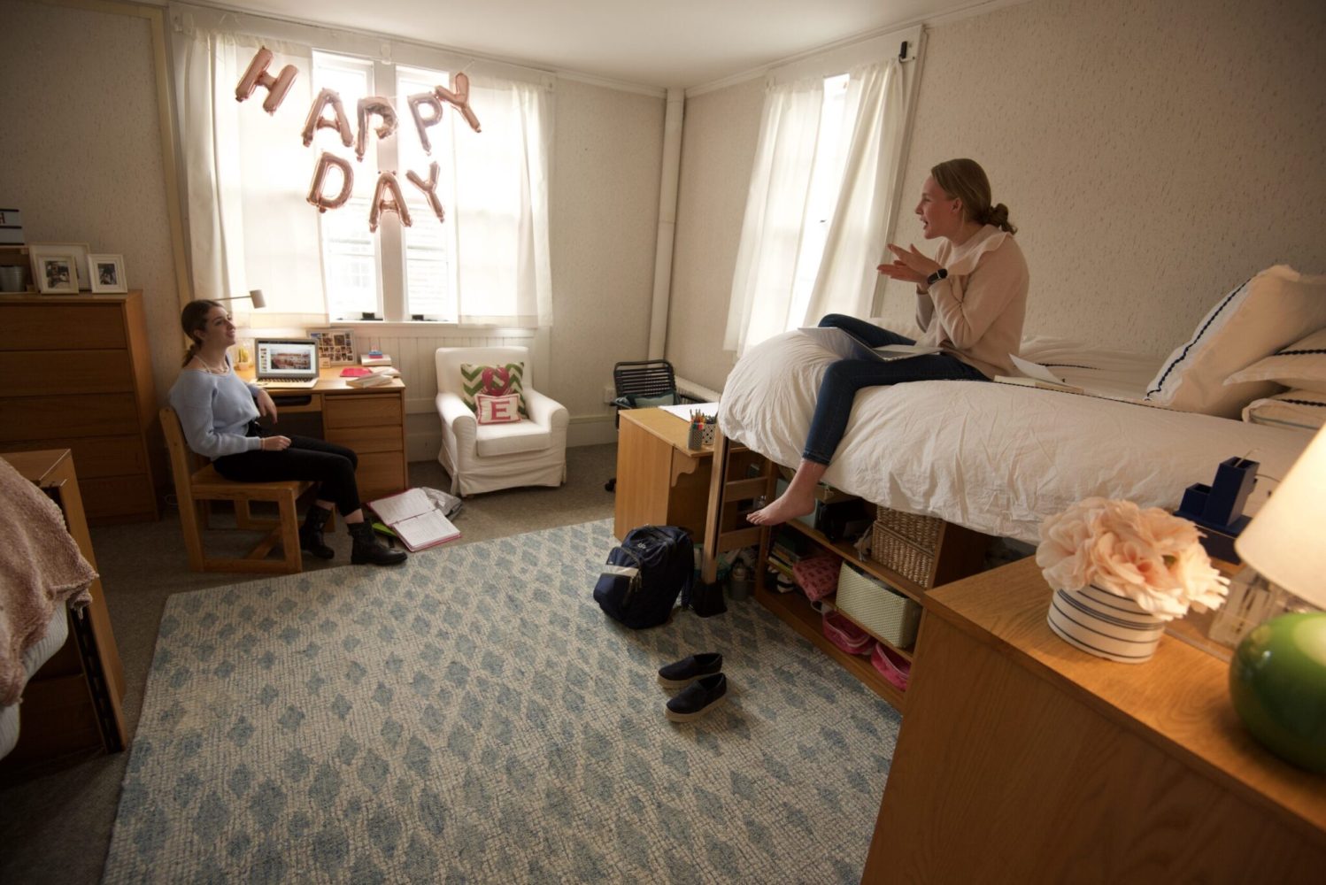 Two women talk in a well-lit dorm room. One sits at a desk with a laptop, the other sits on a bed. HAPPY DAY balloons hang on the window. Room is tidy, with books, decor, and bags.