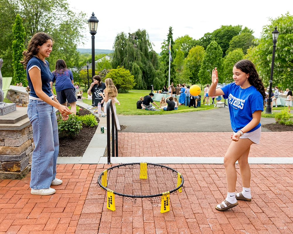 Two girls play Spikeball on a brick path, smiling and active, while others gather and play on a grassy field in the background with trees and a lamppost nearby.