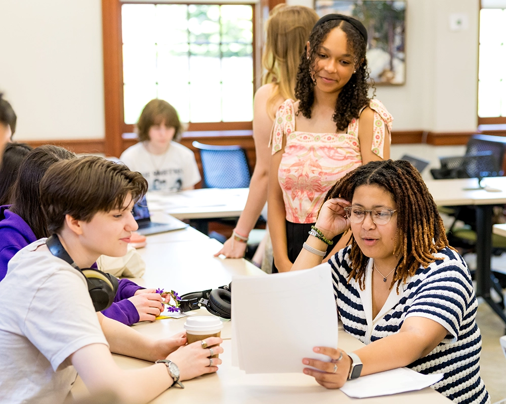 a group of three girls sit around a desk looking at a paper and discussing its contents