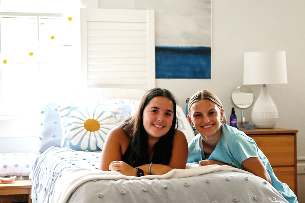 Two young women smile while leaning on a bed with a daisy pillow in a bright, neatly decorated bedroom. A nightstand with a lamp and bottle is in the background.