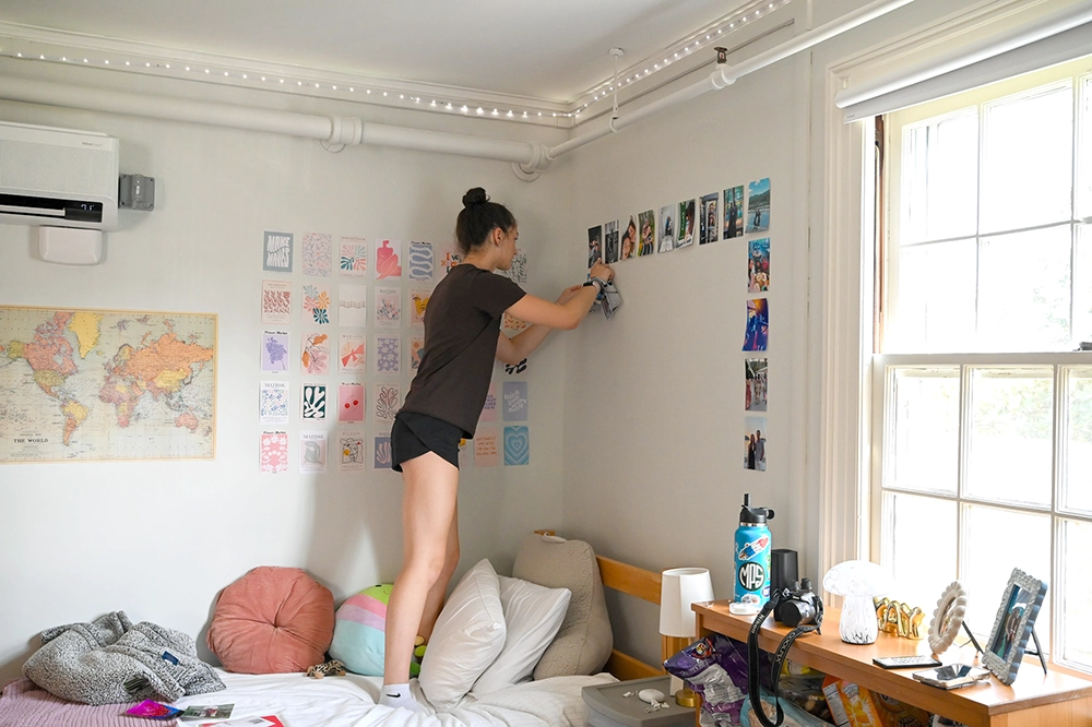 A person stands on a bed, decorating a bedroom wall with photos and art prints. The room has a world map, cozy pillows, a window with sunlight, and string lights along the ceiling.