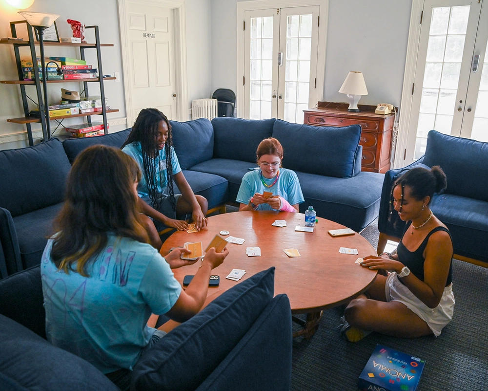 Four people sit around a circular wooden table in a cozy living room, playing a card game. Board games are on a shelf and the floor nearby, and sunlight streams in through glass doors.