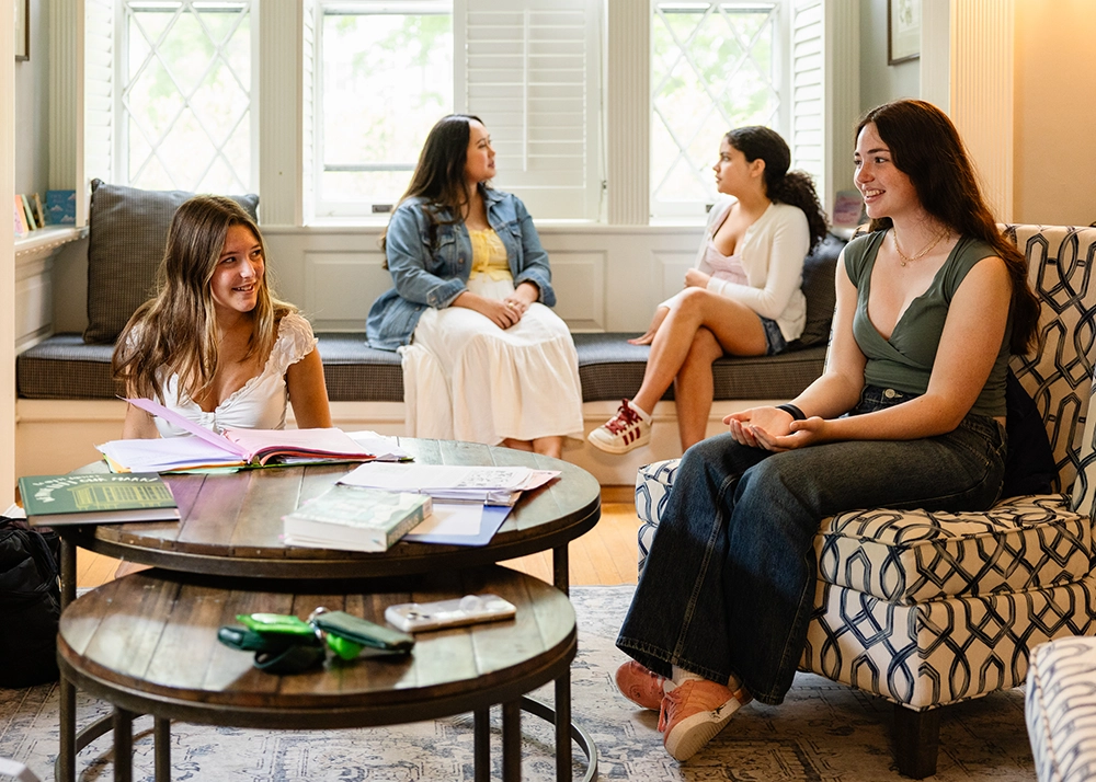 Four young women sit and chat in a cozy living room. Two are at a round table with open books and notebooks, while two more sit by a window bench, engaged in conversation.