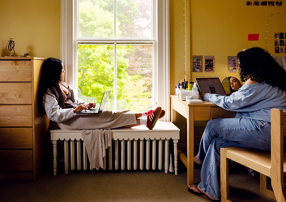 Two people work on laptops in a cozy, yellow room. One sits on a window bench in red sneakers, while the other sits at a desk. A sunny window and wall photos create a warm, relaxed atmosphere.
