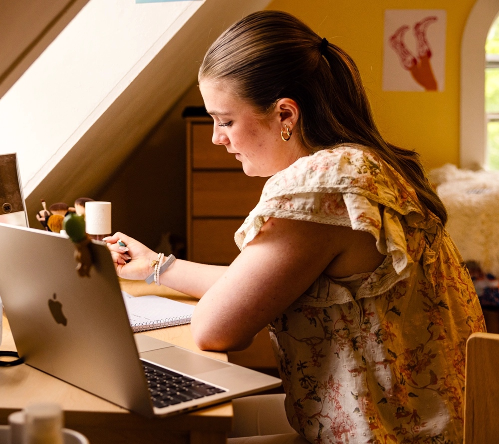 A young woman sits at a desk in a cozy room, writing in a notebook beside an open laptop. She wears a floral blouse and has her hair tied back. Sunlight enters through a nearby window, creating a warm atmosphere.