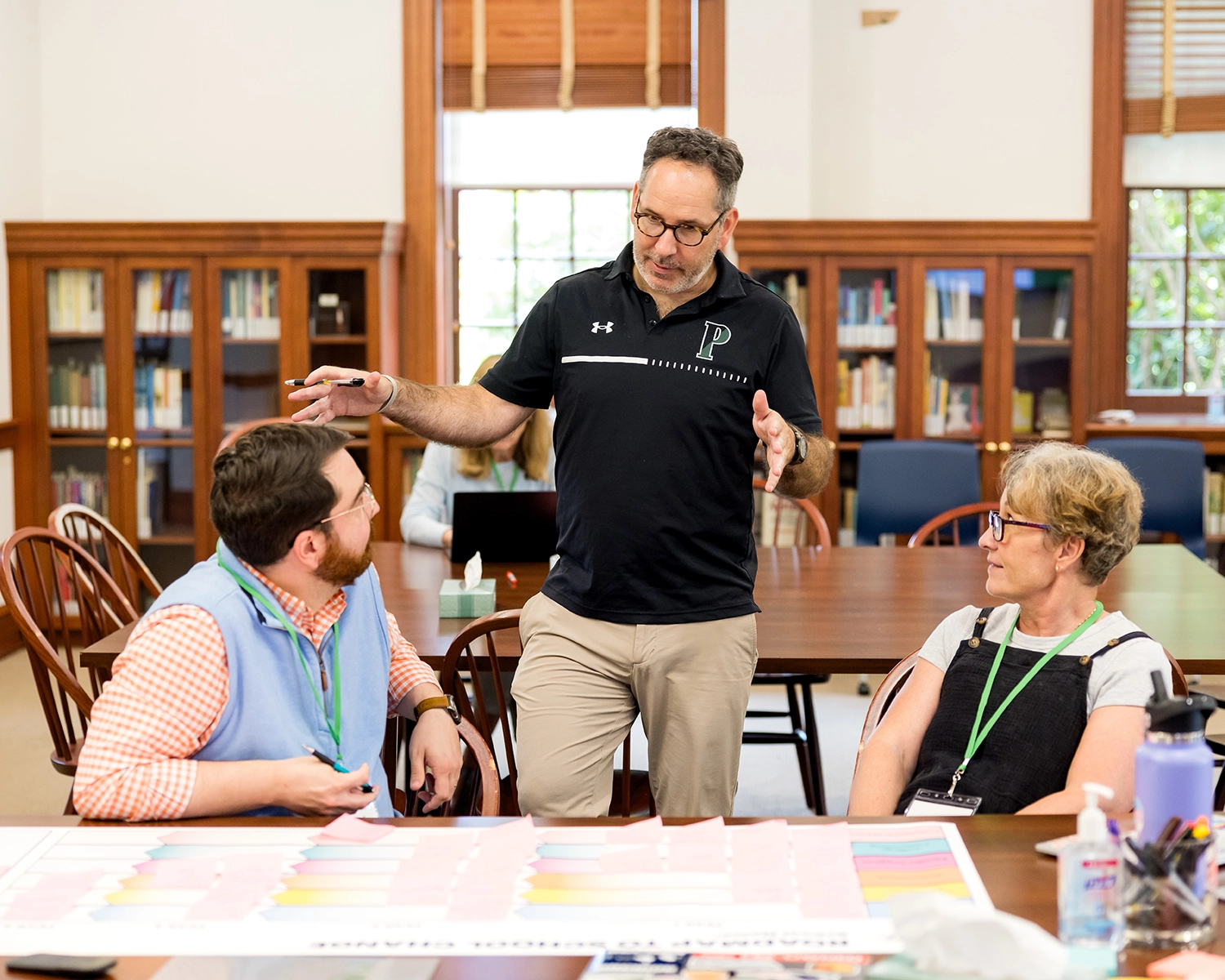Three adults talk around a table in a library or classroom, with bookshelves in the background. One man stands gesturing, while the others sit and listen. A colorful chart is on the table in front of them.