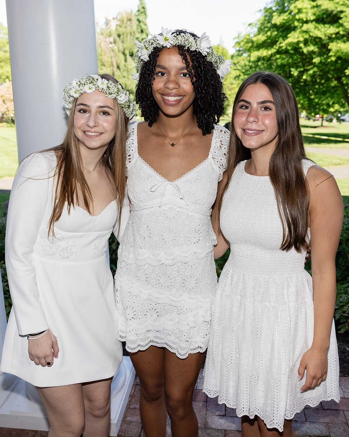 Three young women wearing white dresses and floral crowns stand together, smiling, outdoors on a sunny day with greenery in the background.