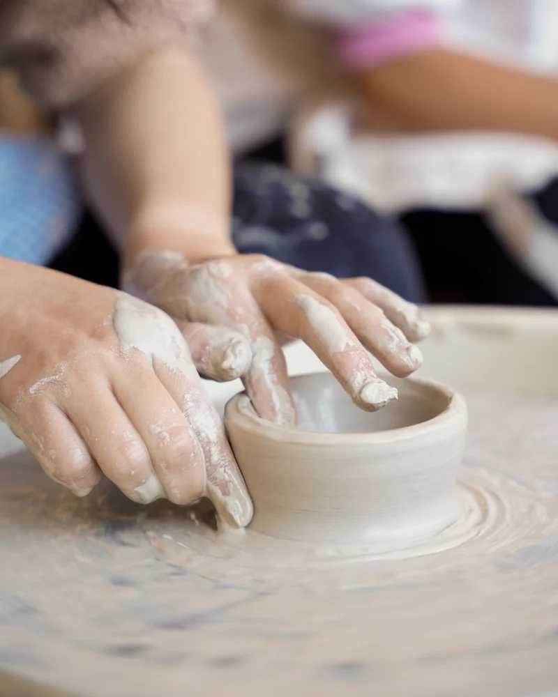 Close-up of two pairs of hands shaping wet clay on a pottery wheel, with fingers covered in clay, creating a small bowl.