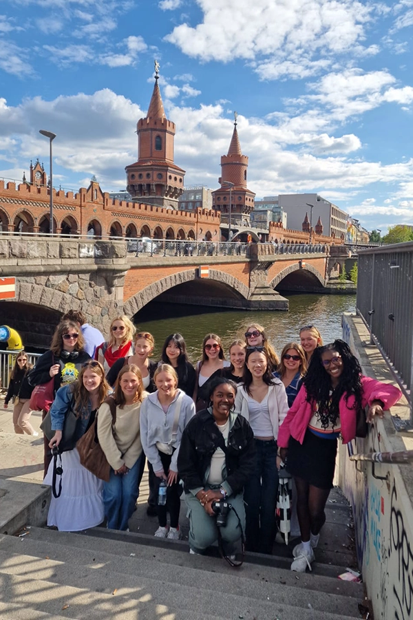 A group of people posing and smiling on steps near a river, with the Oberbaum Bridge and towers in the background under a blue sky with clouds.
