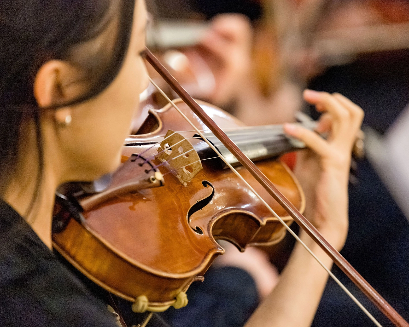 A person plays a violin with focus, holding the instrument with their left hand and using a bow in their right hand. The background shows blurred hands, suggesting other musicians nearby.