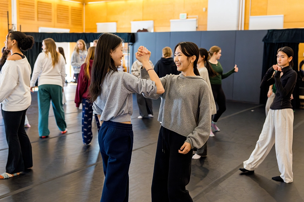 A group of people practice dance in a studio. Two women in the foreground smile and hold each others hands, while others talk or stretch in the background. The setting is casual and friendly.