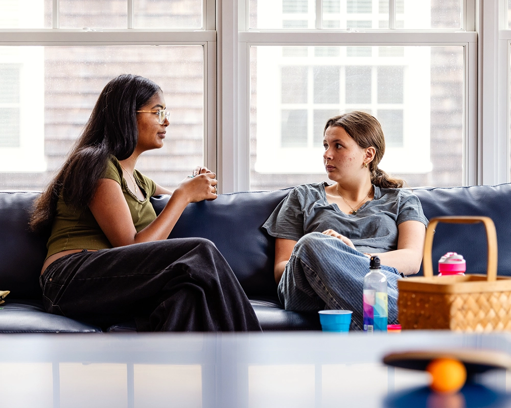 Two women sit on a dark blue couch having a conversation. One has long dark hair and glasses; the other has light brown hair in a ponytail. Sunlit windows and various items are visible in the background and foreground.