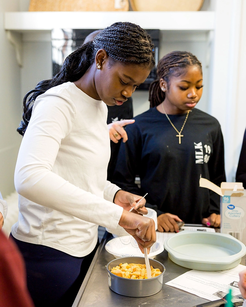 Two young women prepare food together in a kitchen. One stirs ingredients in a bowl, while the other stands nearby watching. Kitchen supplies are visible on the counter.