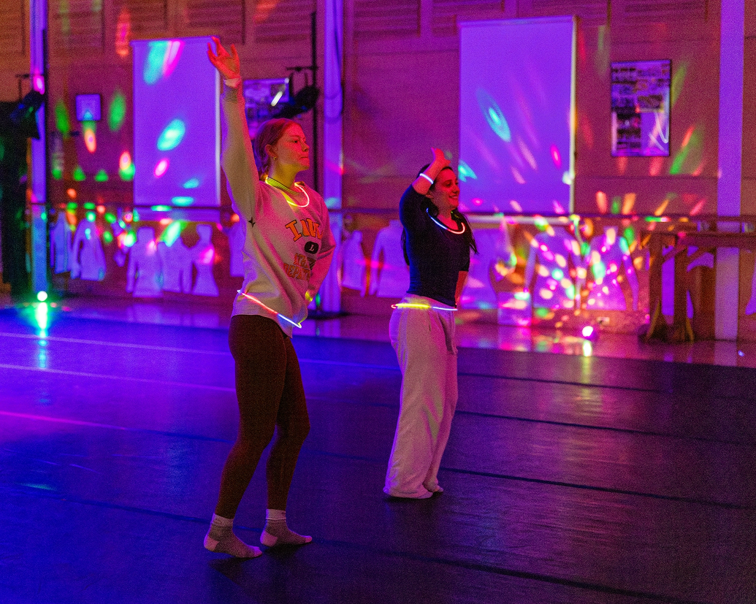Two people dance in a dimly lit room with colorful, swirling lights projected on the walls and floor. Both have one arm raised, and wear casual clothing. The atmosphere appears lively and energetic.