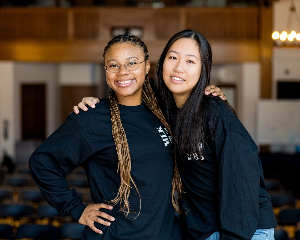 Two young women smiling, standing close with arms around each other, wearing black long-sleeve shirts. They are indoors, with blurred chairs and warm lighting in the background.