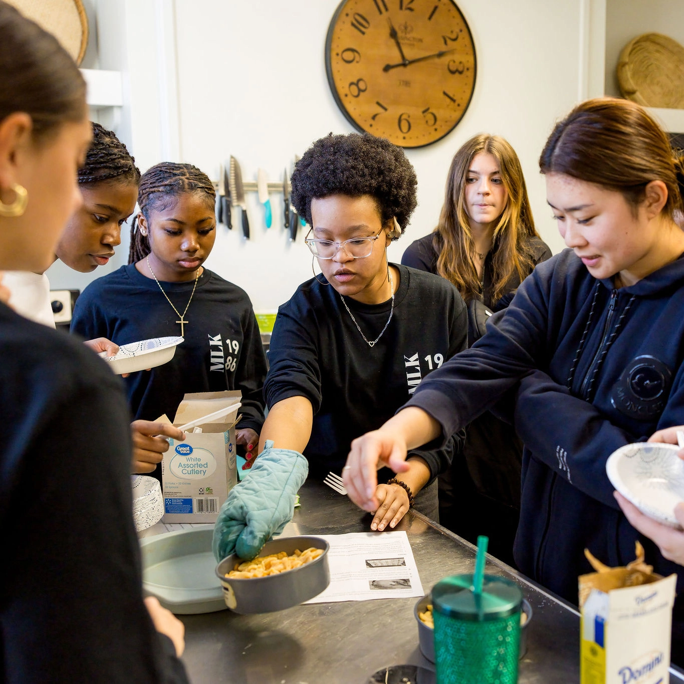 A group of young women gather around a kitchen counter, preparing food together. One person wears an oven mitt and places a dish in a pan. Various ingredients and utensils are on the countertop.
